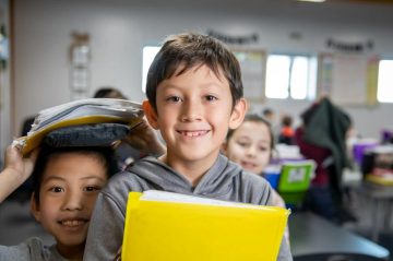 Elementary school boy posing for a photo in a classroom, holding a yellow folder. Another boy is next to him, posing with his folder on top of his head.